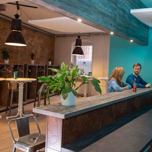 Zwei Menschen sitzen im Gespräch an der Bar. Pflanzen stehen verteilt im Raum. Im Hintergrund stehen weiße Stehtische und Barhocker. An der Wand stehen Garderoben aus grauem Holz.