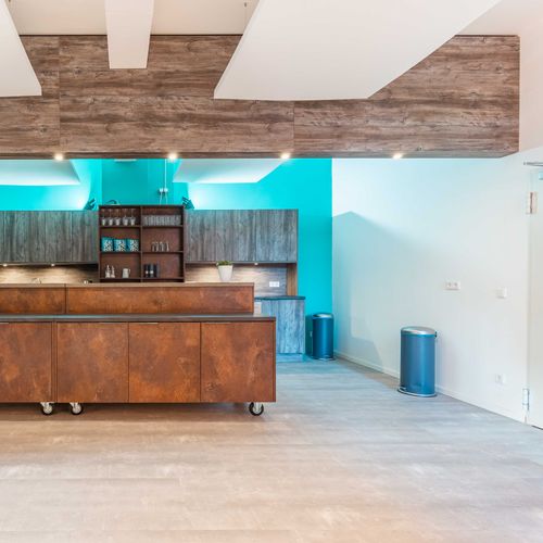  Eine Küchenzeile inklusive beweglicher Kochinsel-Bar mit Elementen aus Holz- und industrieller Messing-Optik stehen unter hellen Oberlichtern. An der Decke sind schallabsorbierende Deckensegel aus weißem Schaumstoff angebracht.